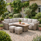 Corner seating arrangement and two cushioned stools on gravel beside a stone house, with flowers and a fruit bowl on the slatted table and a throw over the seat.