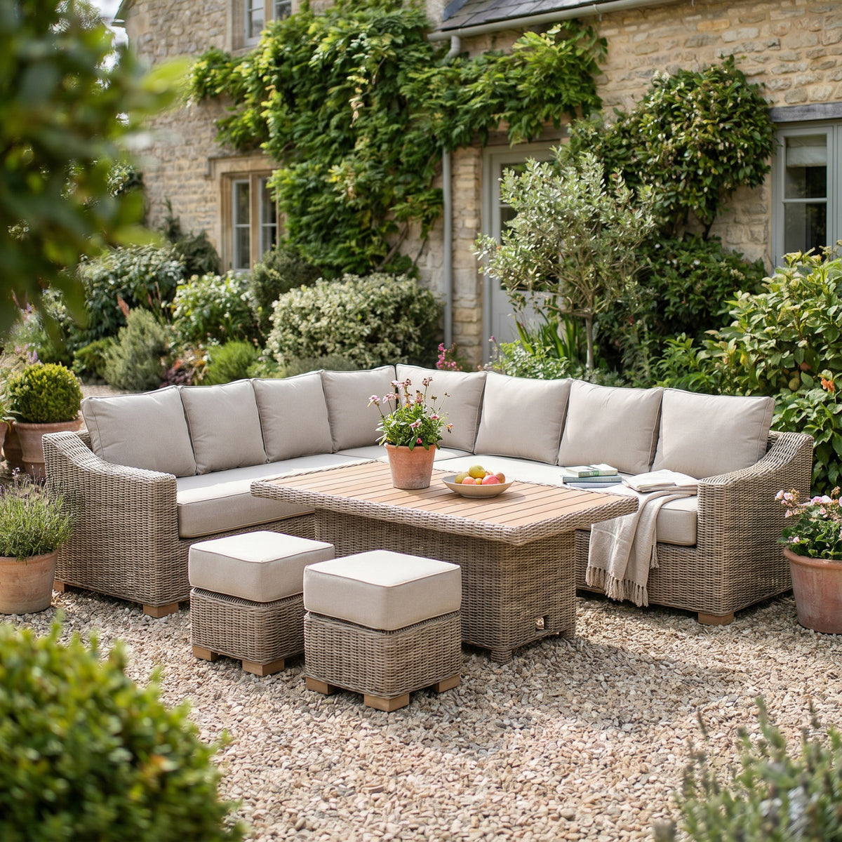 Corner seating arrangement and two cushioned stools on gravel beside a stone house, with flowers and a fruit bowl on the slatted table and a throw over the seat.
