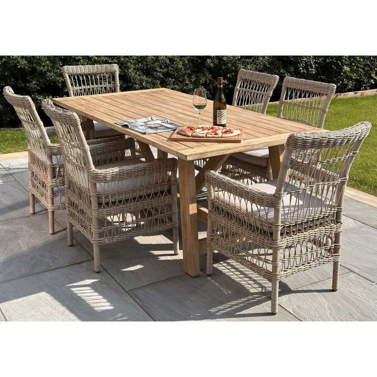 Brown rectangular slatted dining table on a paved patio, surrounded by woven chairs with pale cushions and styled with pizza, a wine bottle and a glass.