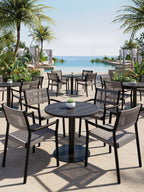 Black round bistro tables with grey woven chairs and small potted plants on a poolside terrace overlooking the sea.