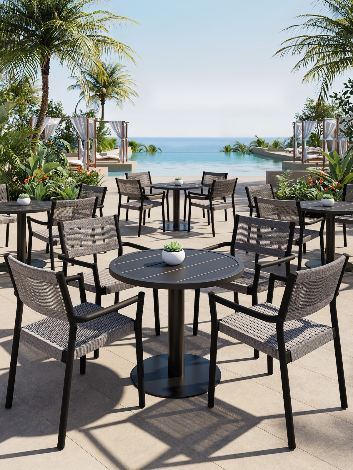 Black round bistro tables with grey woven chairs and small potted plants on a poolside terrace overlooking the sea.