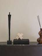 Black figure sculpture on a dark sideboard beside a pale geometric ornament on a black box and an amber glass reed diffuser.