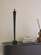Slender black figure sculpture displayed on a dark sideboard beside stacked art books and an amber glass vase with black reeds.