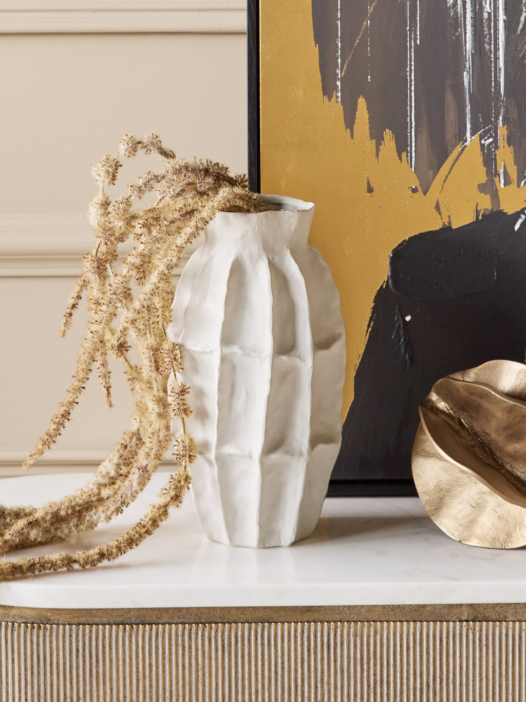 White panelled vase holding trailing dried stems on a pale console top, beside a gold-toned bowl and dark abstract artwork.