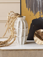 White panelled vase holding trailing dried stems on a pale console top, beside a gold-toned bowl and dark abstract artwork.