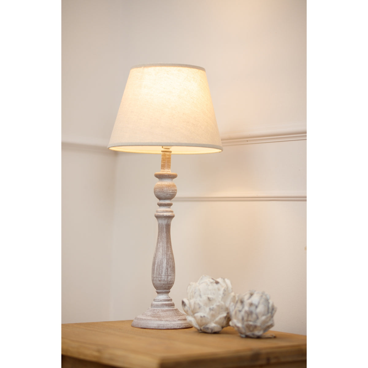 Column table lamp shown illuminated on a wooden console beside two pale artichoke-shaped ornaments against a cream panelled wall.