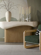 Console table styled with a vase of branches and lit taper candles against a painted wall, with a green armchair in front.