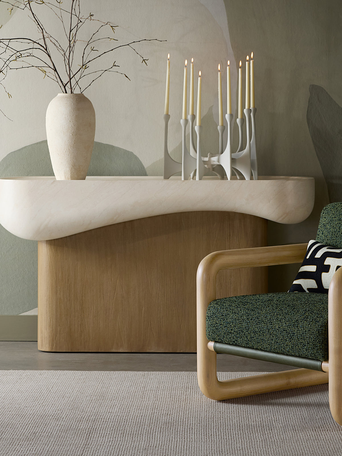 Console table styled with a vase of branches and lit taper candles against a painted wall, with a green armchair in front.