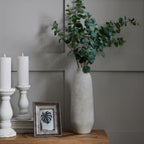 Tall pale grey vase styled with leafy green stems on a wooden console beside white candlesticks and a framed leaf print against a grey panelled wall.