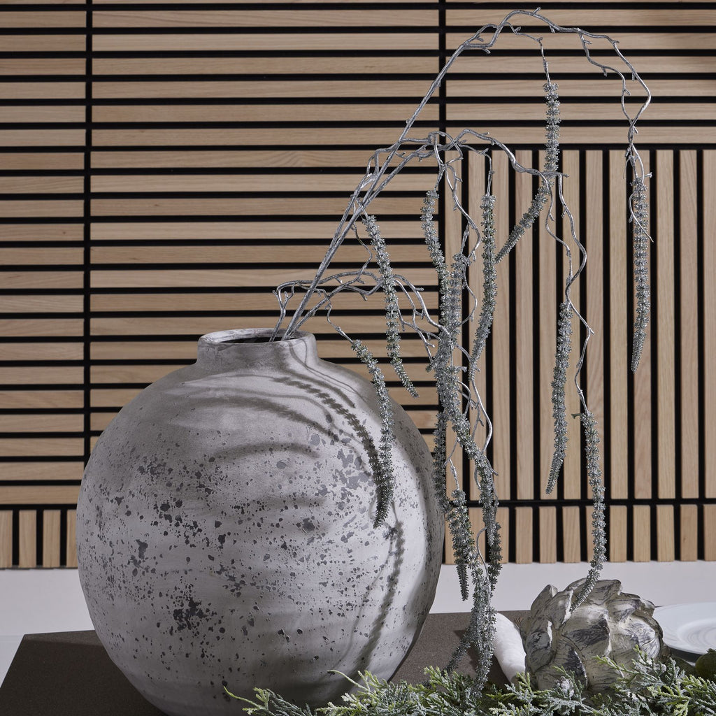 Silver-coloured amaranthus spray with long textured tassels in a rounded grey vase, styled beside greenery and an artichoke-shaped ornament against a slatted wall.