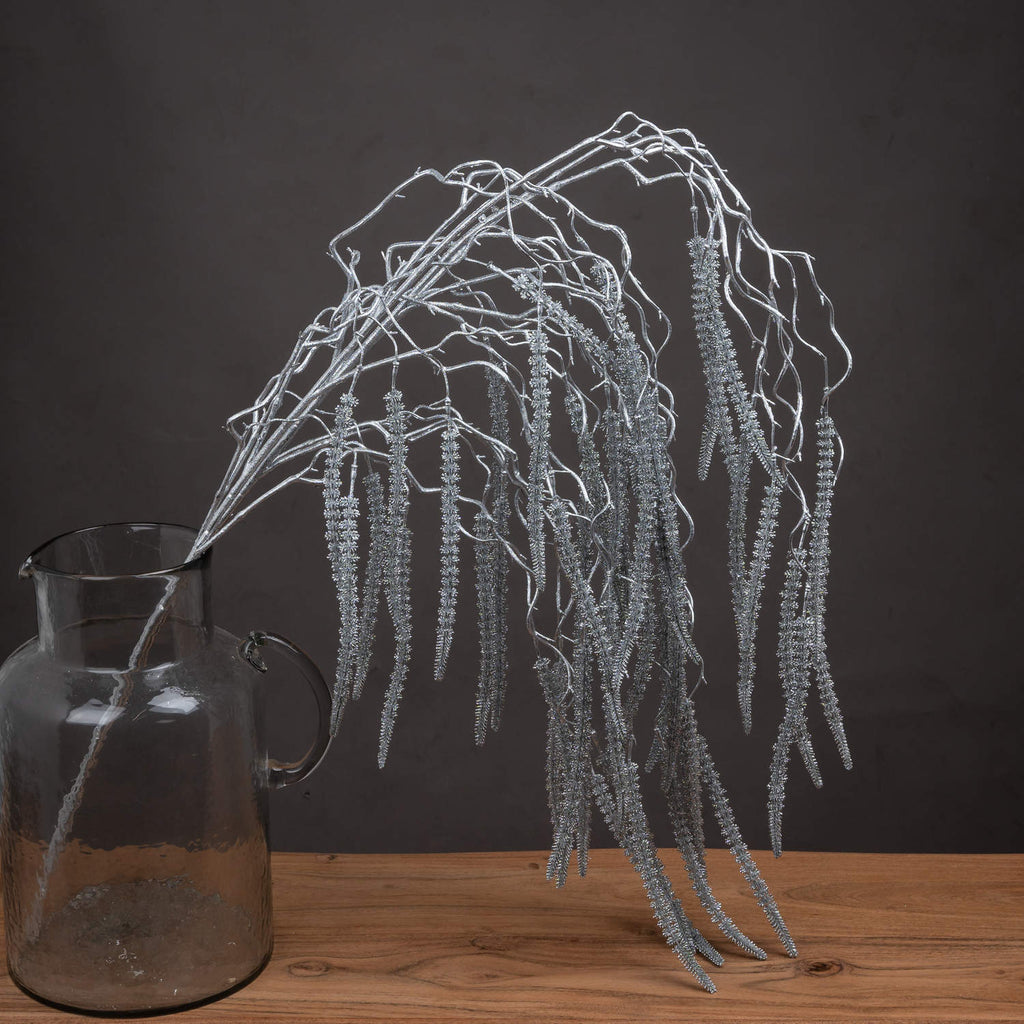 Silver-coloured artificial amaranthus cascading from a smoky glass jug on a wooden surface against a dark grey backdrop.