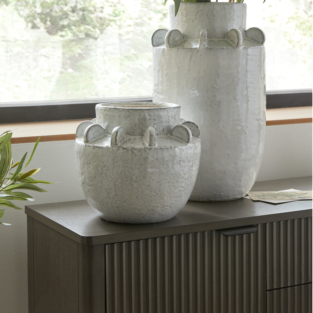 Rounded speckled vase displayed on a dark ribbed cabinet in front of a taller matching vase beside a window.