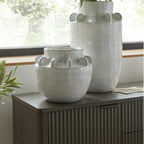 Rounded speckled vase displayed on a dark ribbed cabinet in front of a taller matching vase beside a window.