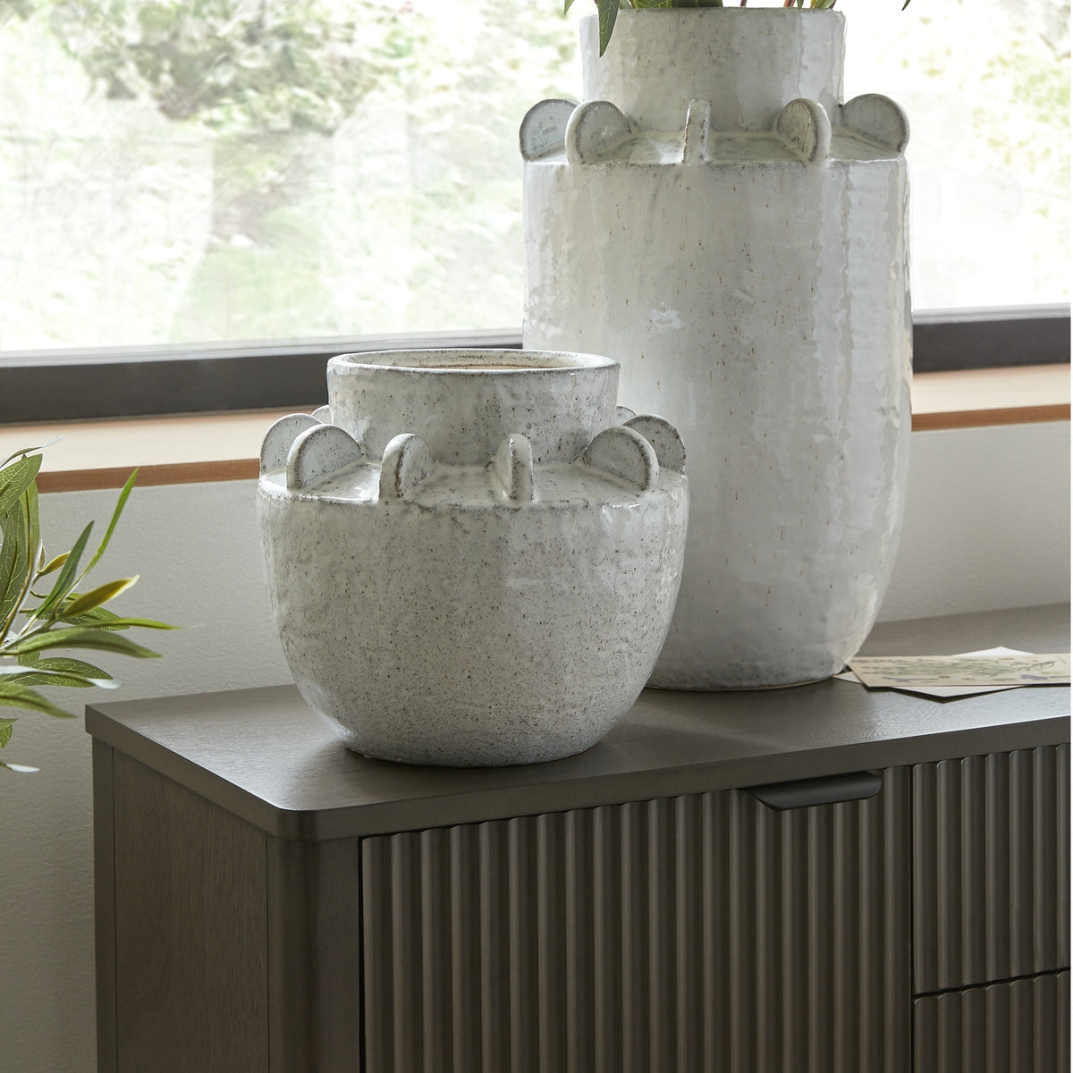 Rounded speckled vase displayed on a dark ribbed cabinet in front of a taller matching vase beside a window.