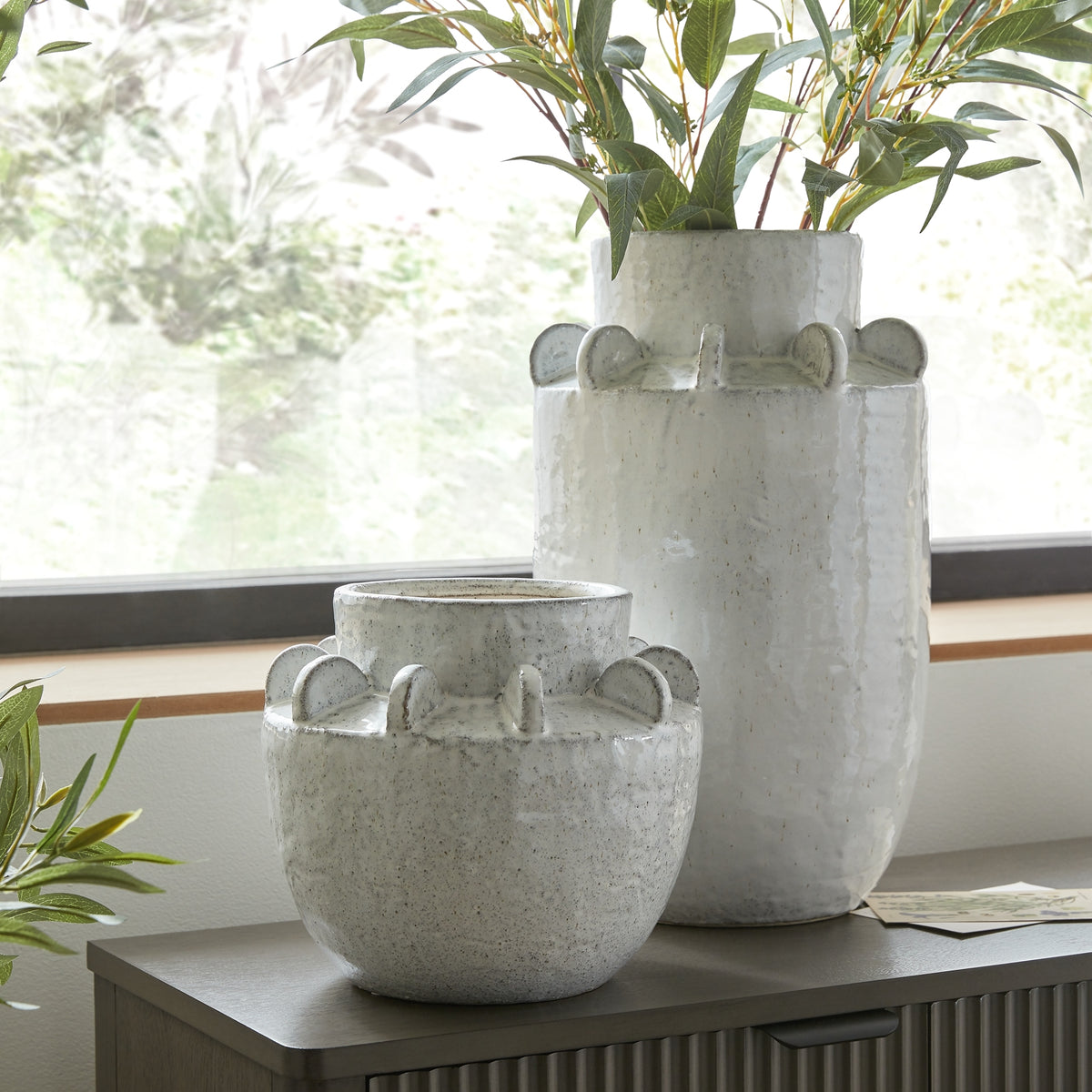 Tall speckled shoulder-finned vase styled with leafy stems beside a shorter rounded vase on a dark cabinet beneath a window.