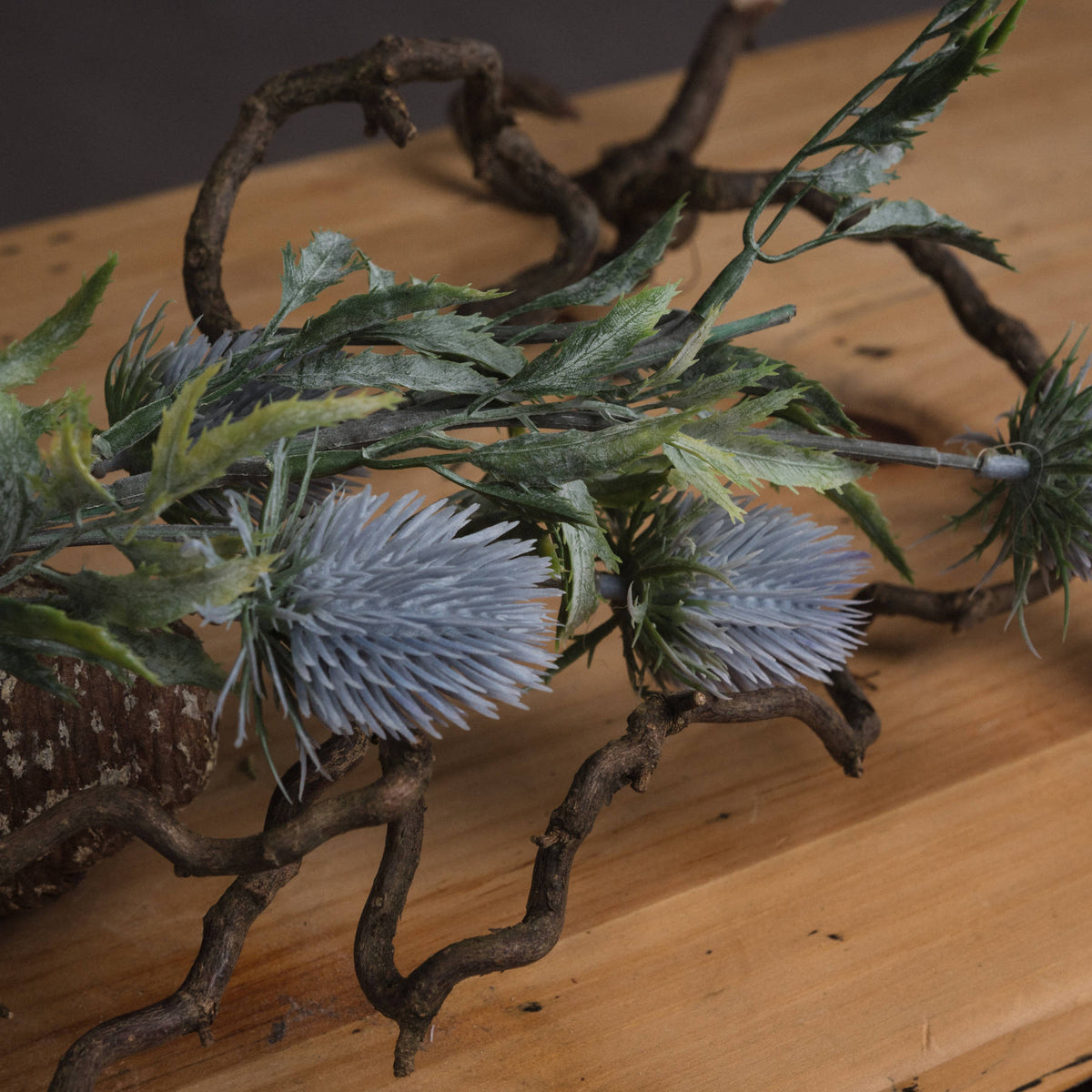 Close-up of spiked grey-blue thistle heads and serrated green-grey leaves laid across twisted branches on a wooden surface.