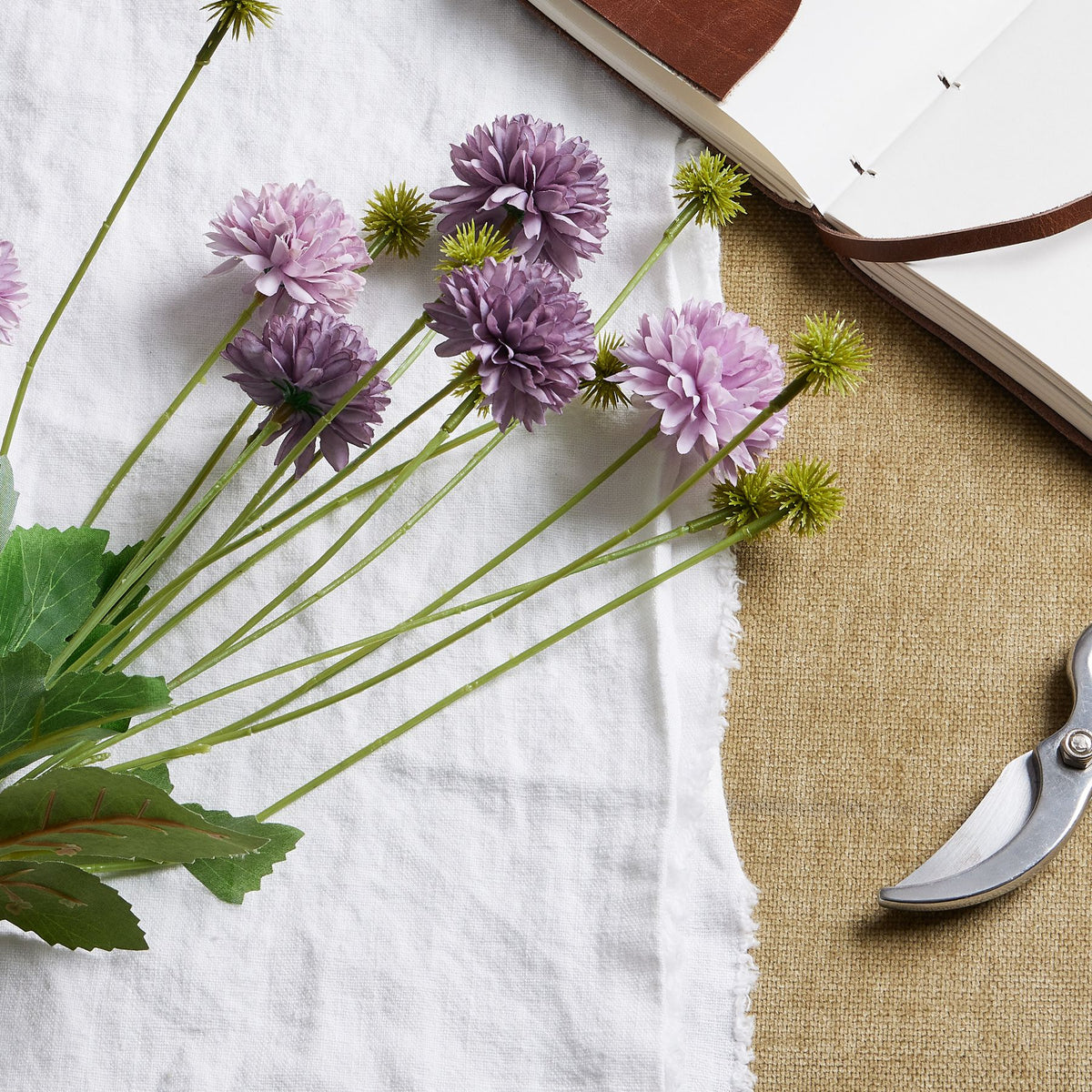 Close view of layered lilac and purple blooms among pointed green buds, laid across white cloth beside an open notebook and part of a pair of secateurs.