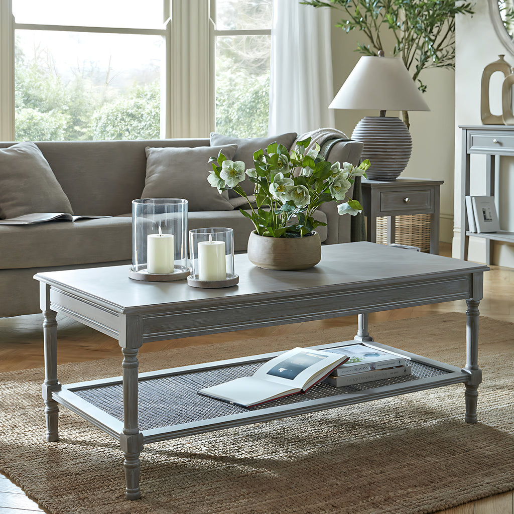 Coffee table in front of a neutral sofa on a textured rug, styled with glass candle holders and white flowers on top and books on the woven shelf.
