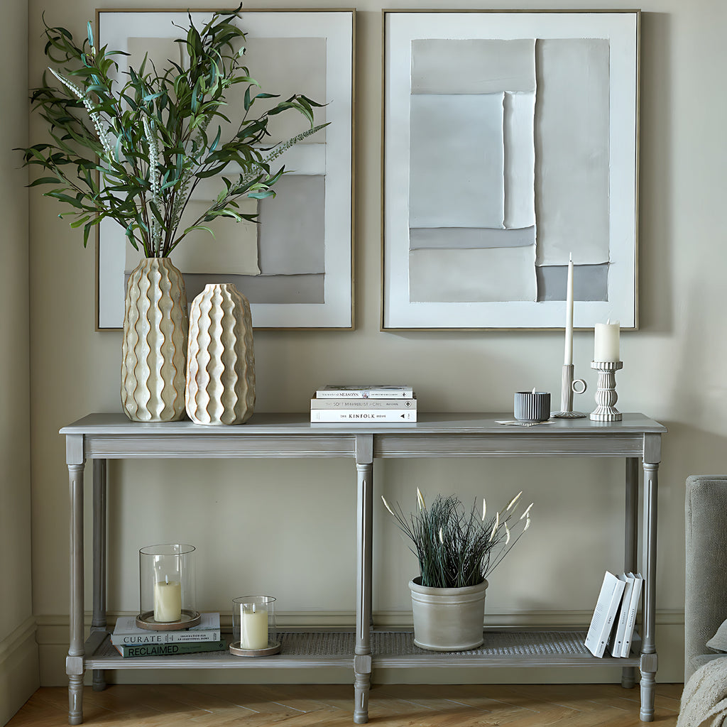 Grey console beneath two pale abstract artworks, styled with tall ceramic vases, books and candleholders, with a potted plant and more books below.
