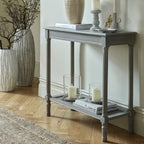 Console table against a pale wall, styled with a ceramic vase, books, candleholders and a dish, with glass candle vessels on the woven shelf and tall floor vases alongside.