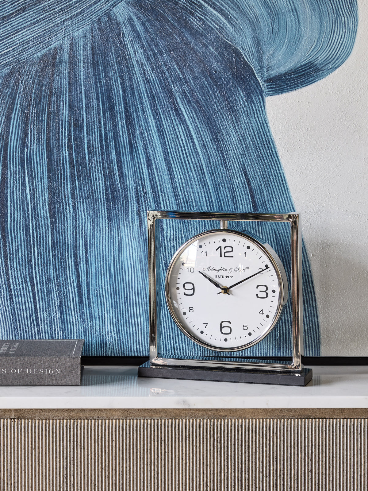 Square frame mantel clock on a pale console top beside a book, with a large blue artwork behind.