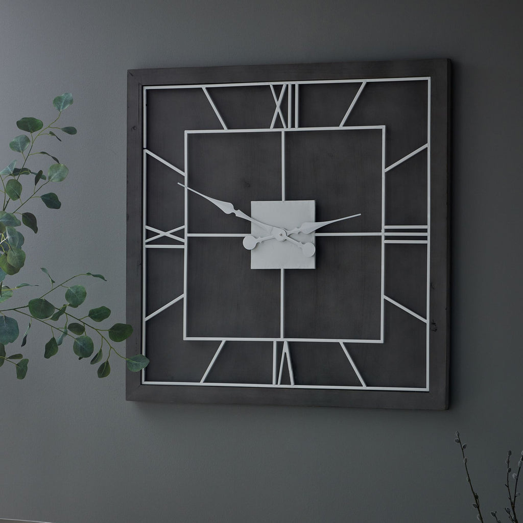 Square grey and white framework clock hanging on a grey wall beside leafy green branches.