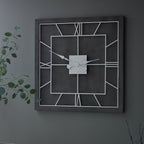 Square grey and white framework clock hanging on a grey wall beside leafy green branches.