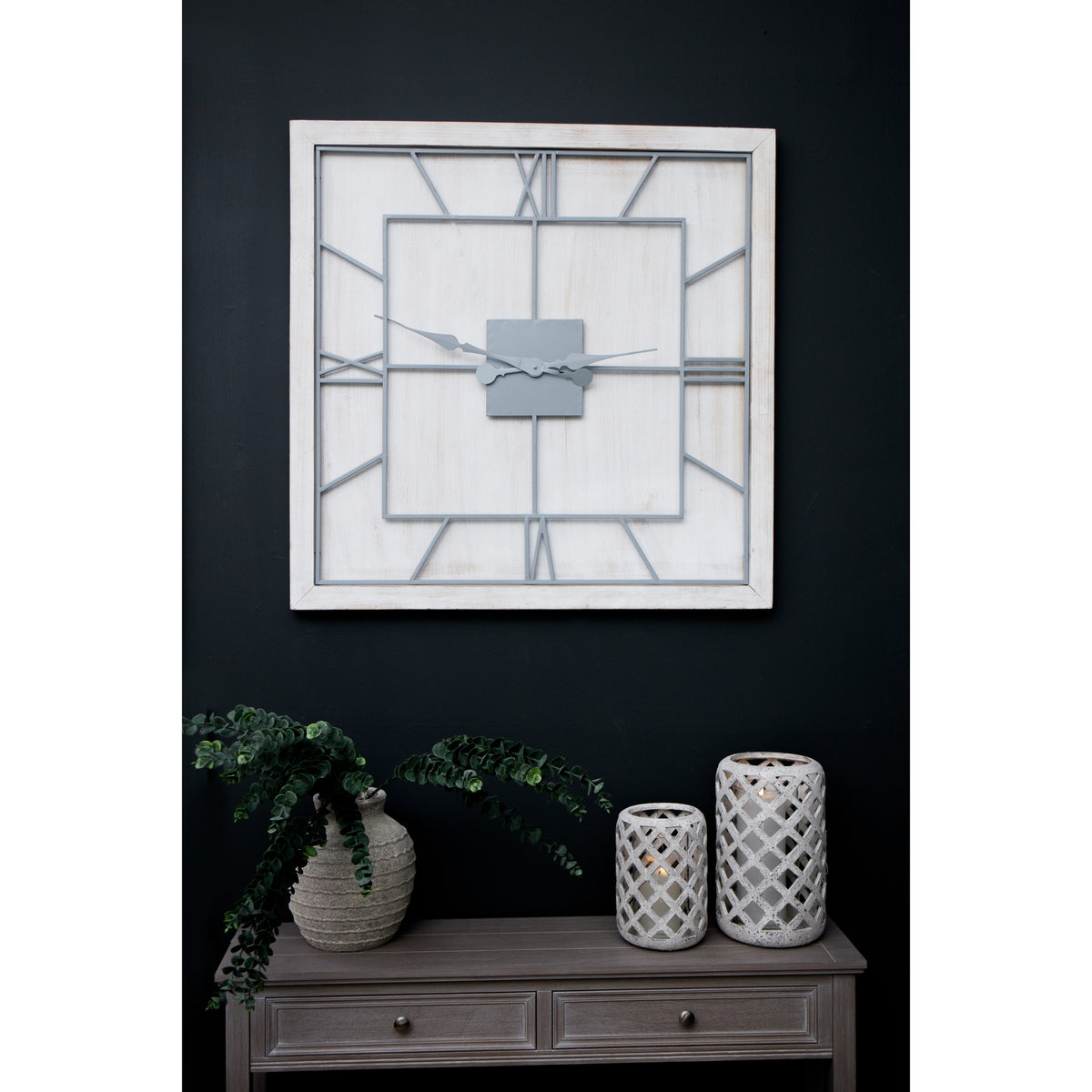 Whitewashed square clock on a dark wall above a grey console, styled with trailing greenery and two pale lattice candle holders.