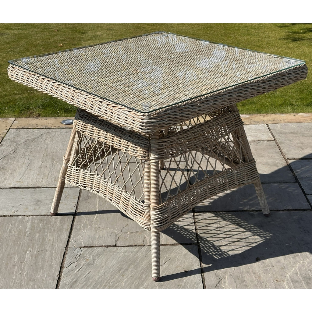 Angled view of a beige square table on stone paving, showing its transparent top over a woven pattern and open crossing-strand panels below.