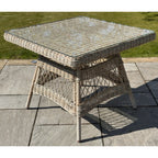 Angled view of a beige square table on stone paving, showing its transparent top over a woven pattern and open crossing-strand panels below.