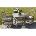 Beige square table staged with cushioned armchairs on a stone patio beside a lawn, with flowers on the transparent tabletop and open patterned chair backs.