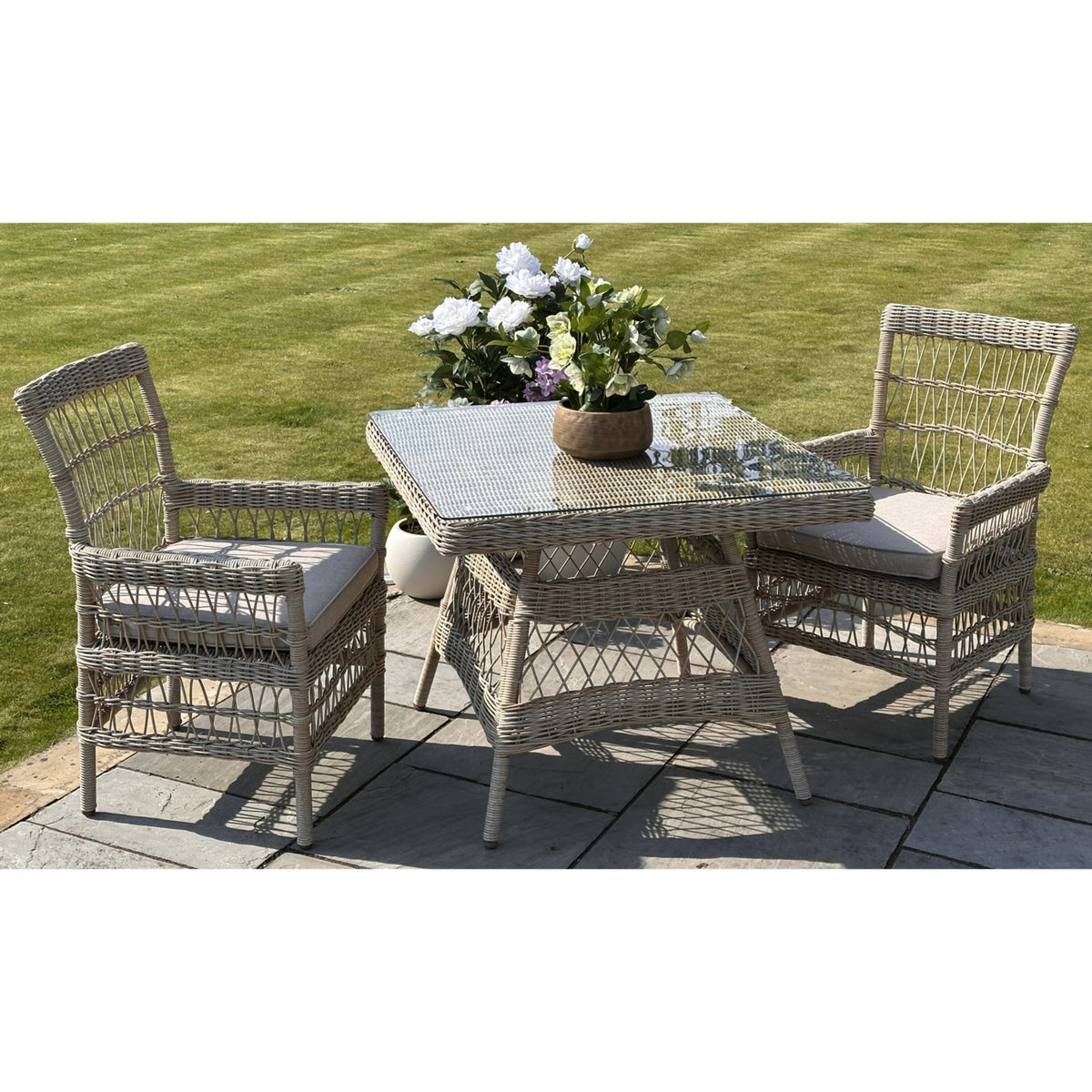 Beige square table staged with cushioned armchairs on a stone patio beside a lawn, with flowers on the transparent tabletop and open patterned chair backs.
