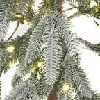 Close-up of green artificial foliage with textured white snow dusting and glowing yellow-toned lights between the branches.