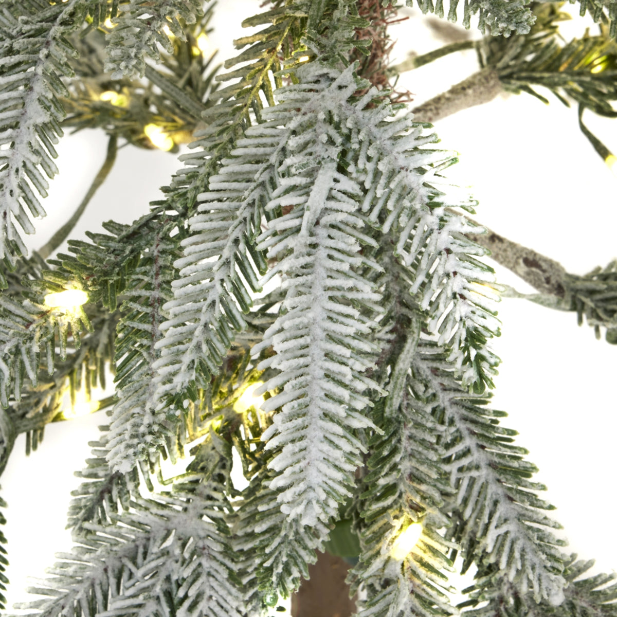 Close-up of green artificial foliage with textured white snow dusting and glowing yellow-toned lights between the branches.
