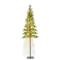 Green artificial woodland Christmas tree shown with warm lights, sparse branches, a long exposed trunk and a dark square base against a white background.