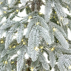 Close-up of green artificial tree branches covered in textured white snow-like dusting, with yellow lights visible between the foliage.