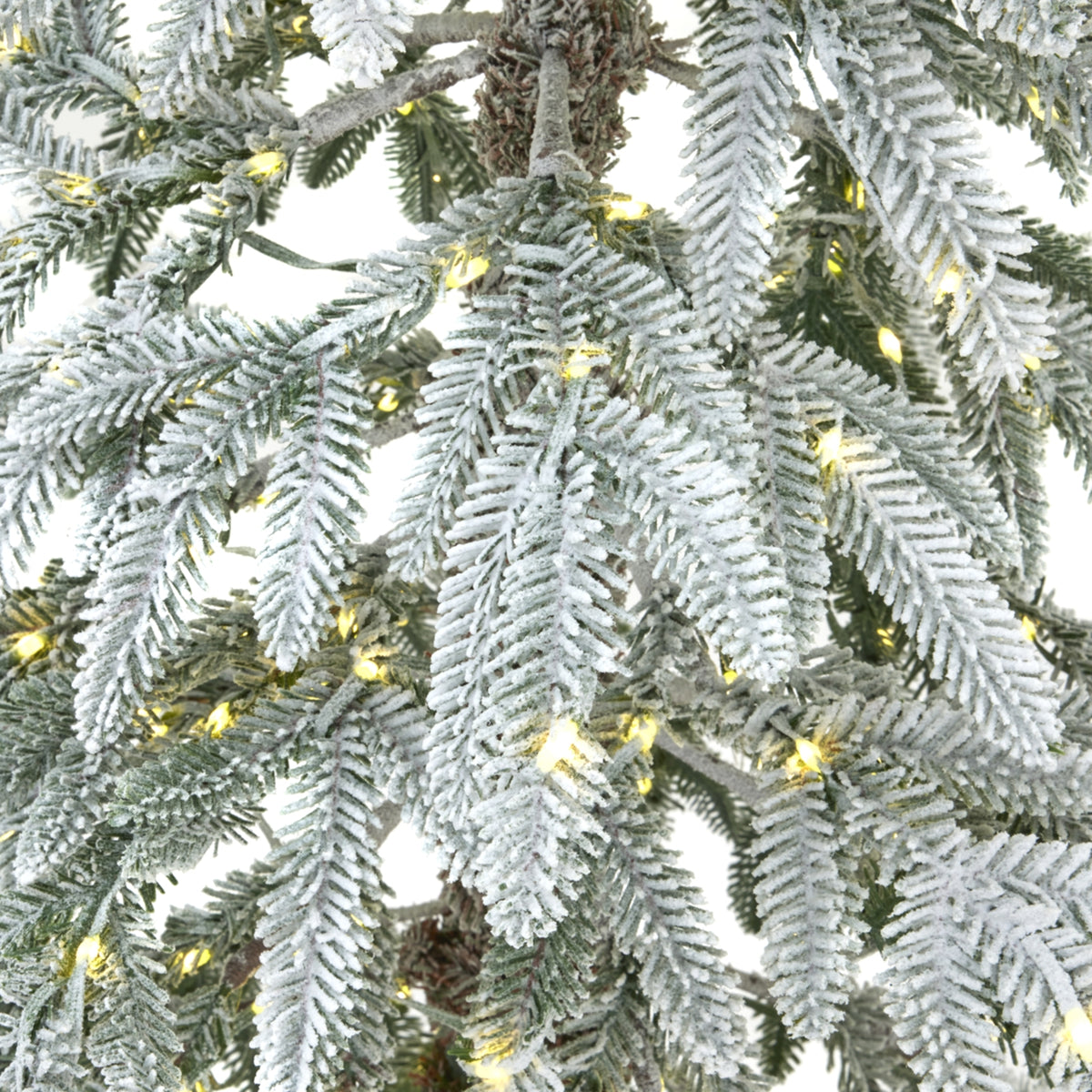 Close-up of green artificial tree branches covered in textured white snow-like dusting, with yellow lights visible between the foliage.