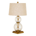 Table lamp with two stacked clear globes around a gold-coloured stem, a stepped square base and a tapered cream textured shade topped by a small finial.