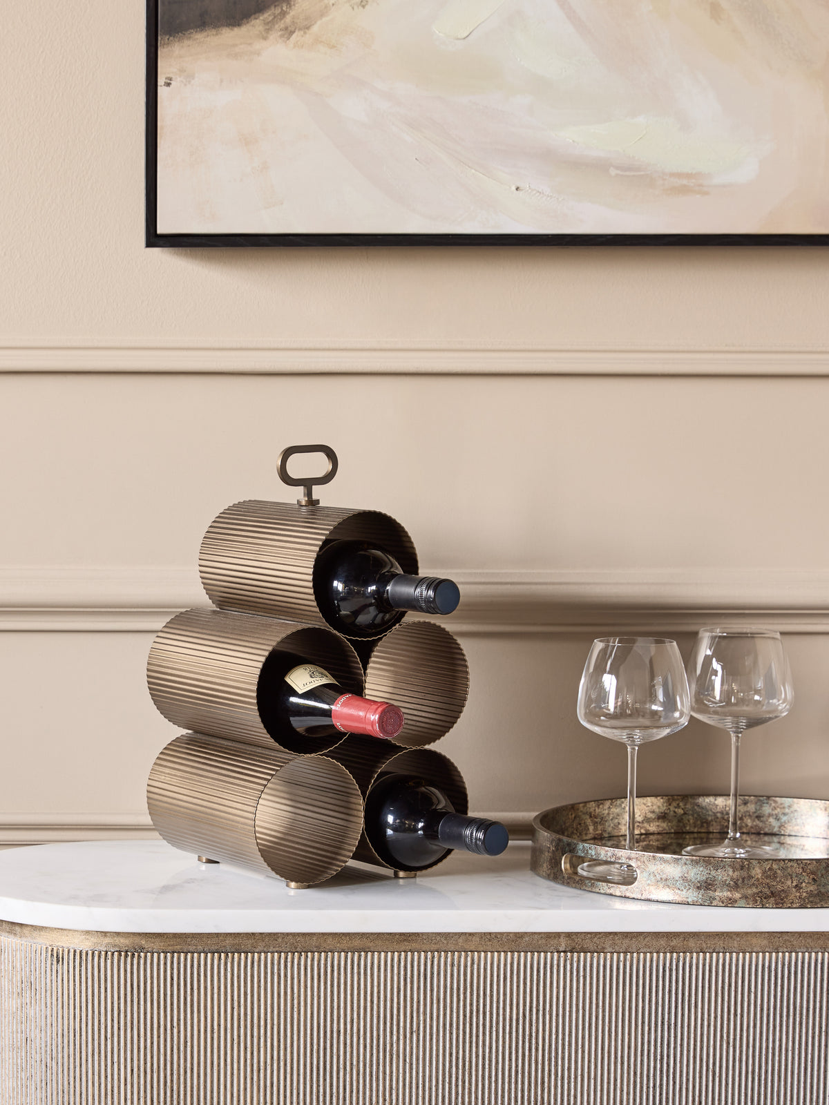 Wine rack holding three bottles on a pale sideboard beside two wine glasses on a round metallic tray.