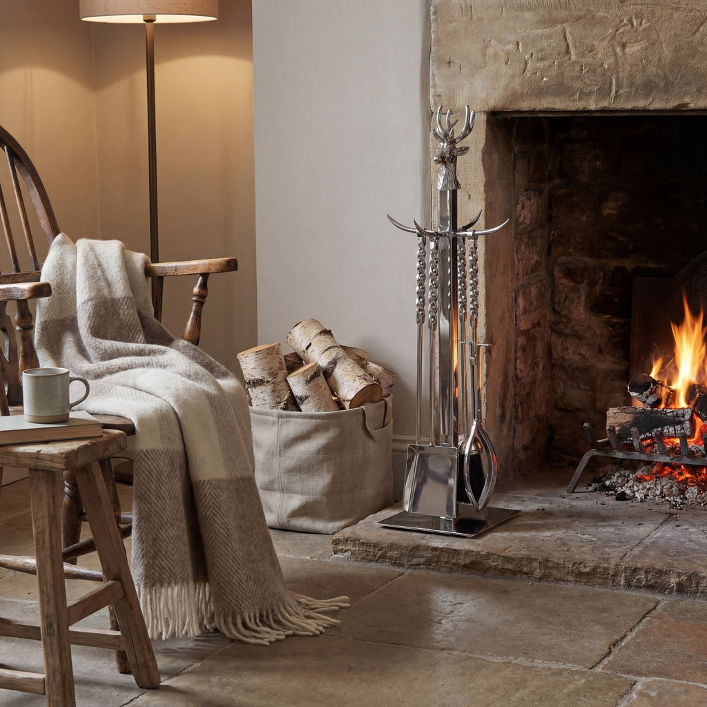 Stag-post fireplace tool set beside a lit stone fireplace, with a bag of logs, wooden chair, pale throw and floor lamp nearby.