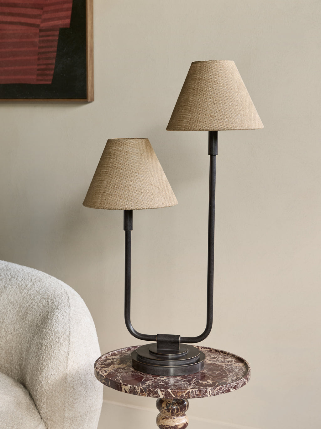 Staggered twin table lamp on a round veined stone side table beside a cream textured armchair, against a pale wall with framed artwork.