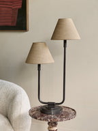 Staggered twin table lamp on a round veined stone side table beside a cream textured armchair, against a pale wall with framed artwork.