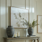 Stemmed Leafscatter artwork on a pale panelled wall above a console styled with dark vases, leafy stems, candles and stacked books.