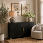 Black sideboard styled beneath two botanical prints with a lamp and flower-filled vase on top, beside a pale armchair and a potted tree.