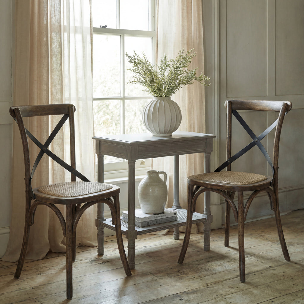 Two Strapcross Dining Chairs beside a pale table with a white vase of foliage, styled in front of a window with light curtains.