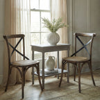 Two Strapcross Dining Chairs beside a pale table with a white vase of foliage, styled in front of a window with light curtains.