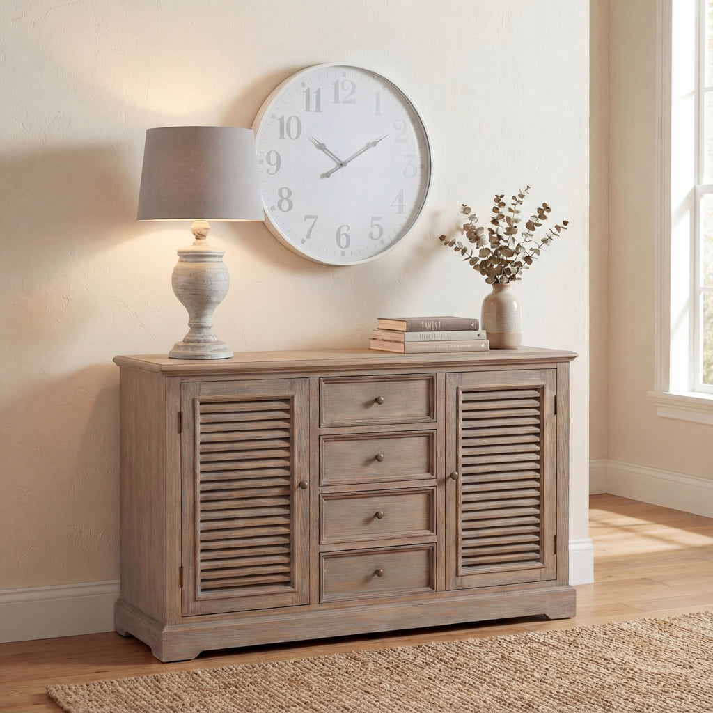 Illuminated urn table lamp on a wooden sideboard beneath a large round wall clock, with stacked books and a vase of leafy stems nearby.