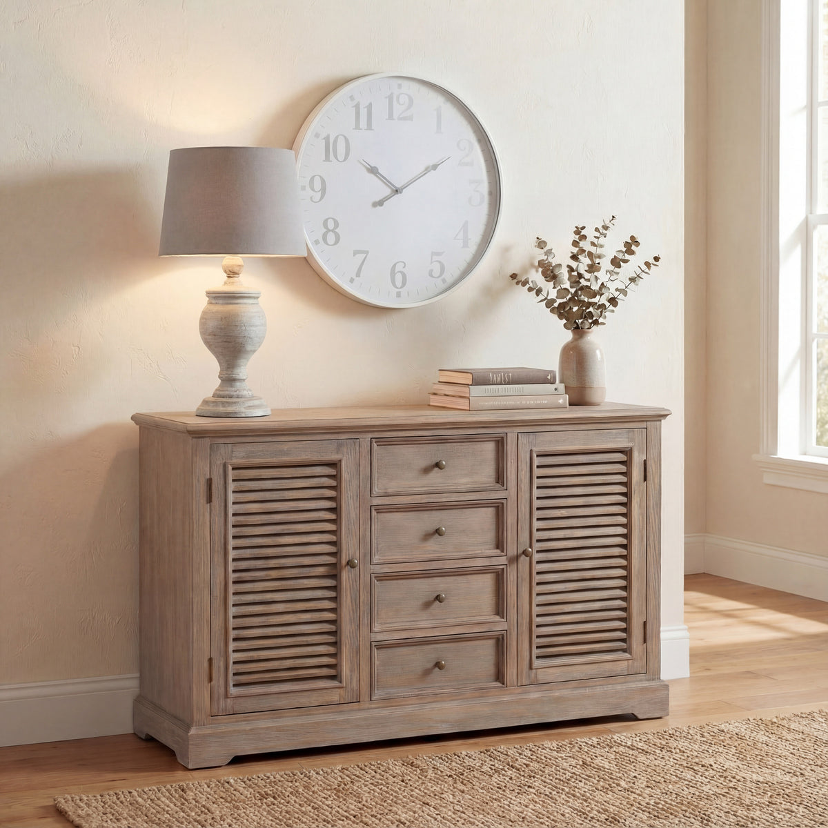 Illuminated urn table lamp on a wooden sideboard beneath a large round wall clock, with stacked books and a vase of leafy stems nearby.