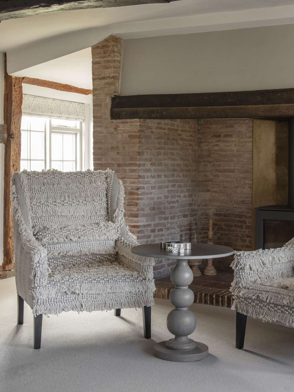 Dice and striped tray on a grey pedestal table between textured armchairs beside an exposed brick fireplace.