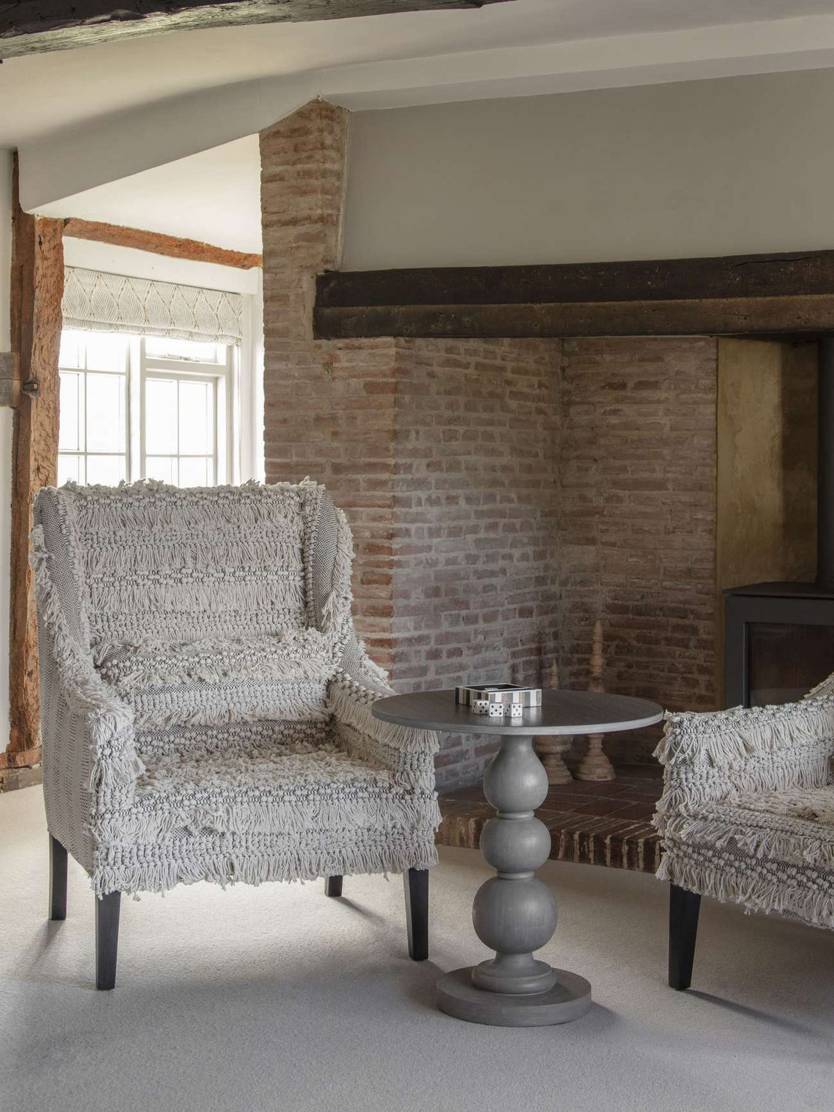 Dice and striped tray on a grey pedestal table between textured armchairs beside an exposed brick fireplace.